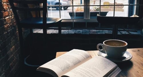 Cozy café scene with an open book and a steaming coffee cup on a wooden table near a window