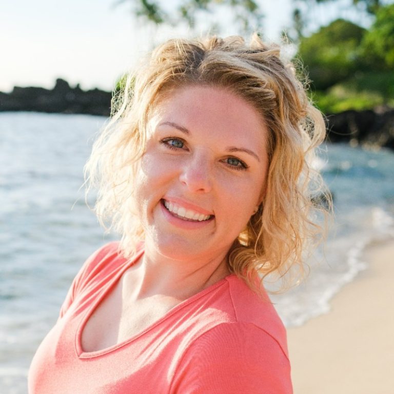 Smiling Kourtney with curly blonde hair on a sunny beach with waves and greenery in the background