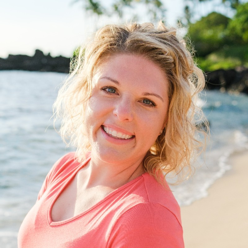 Smiling Kourtney with curly blonde hair on a sunny beach with waves and greenery in the background