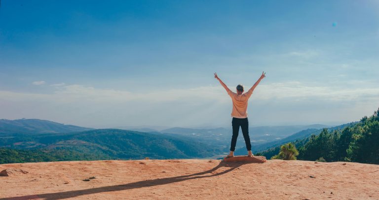 Person standing on hilltop with arms raised, overlooking scenic mountain landscape under clear blue sky