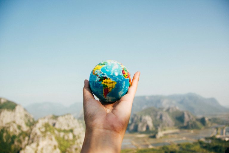 Hand holding a small globe against scenic mountain landscape under clear blue sky