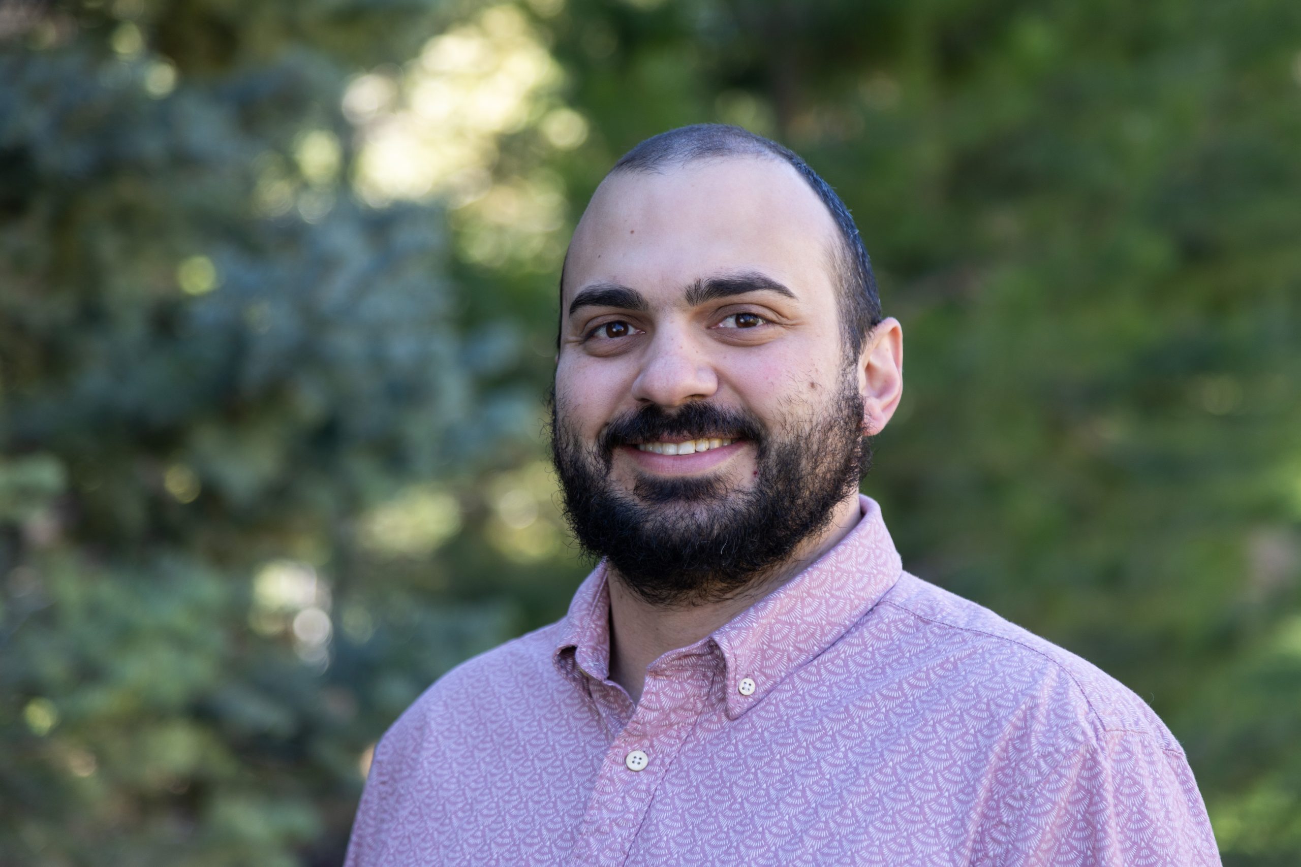 Smiling man with a beard wearing a pink shirt outdoors in a lush green setting