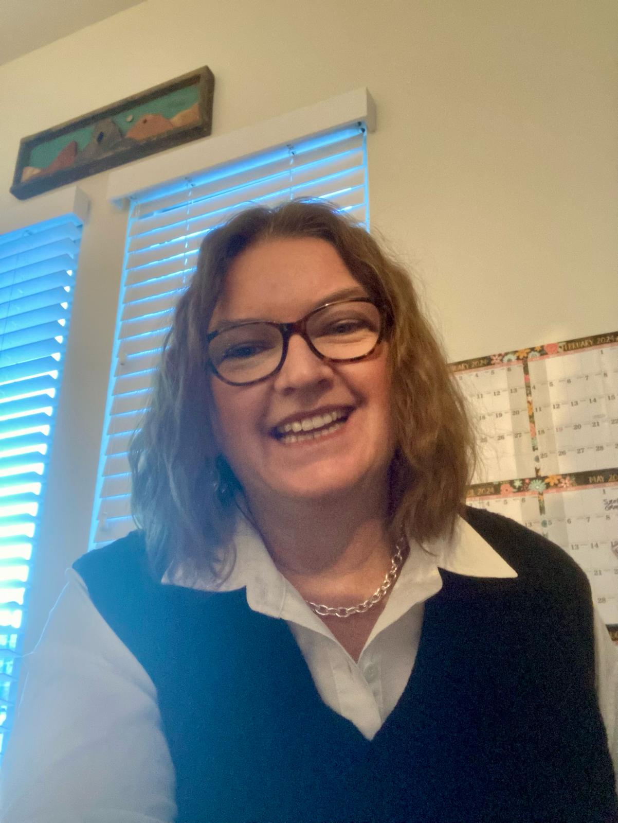 Smiling person with glasses and brown hair wearing a black vest, in a room with calendars and blinds