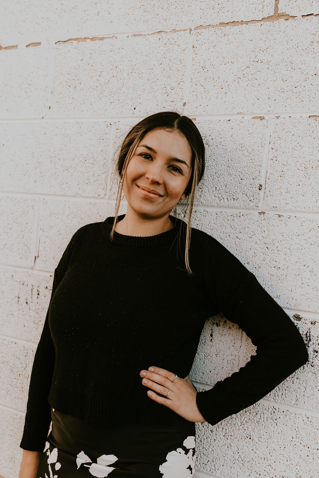 Smiling Tori in black sweater leaning against a white brick wall with hand on hip
