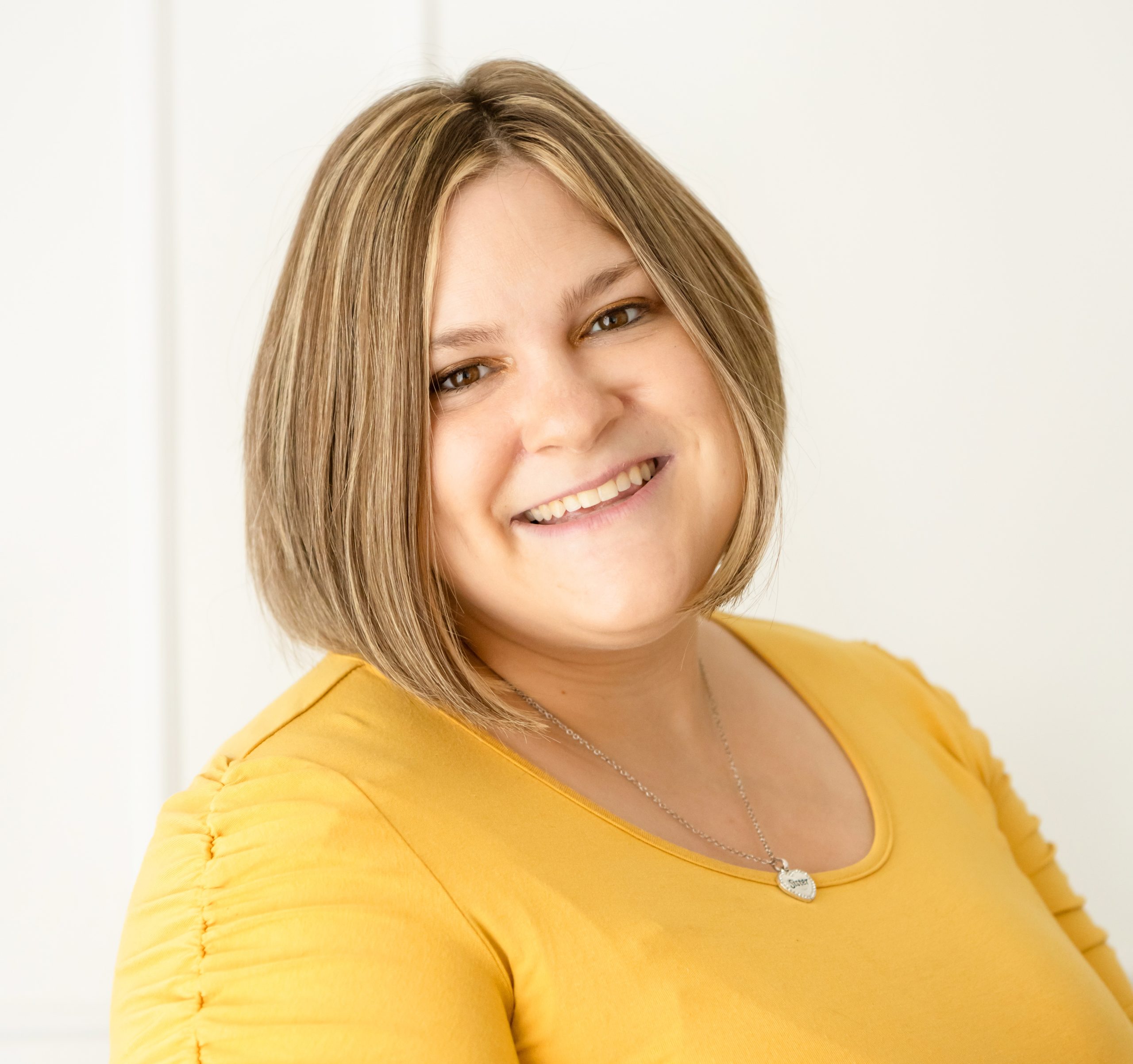 Smiling person in a yellow shirt with short hair, wearing a necklace against a light background