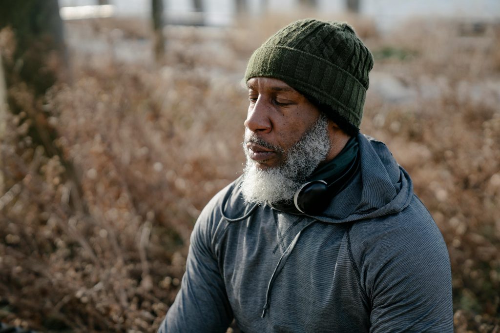 Bearded senior male wearing warm clothes with headphones on neck sitting with closed eyes in nature