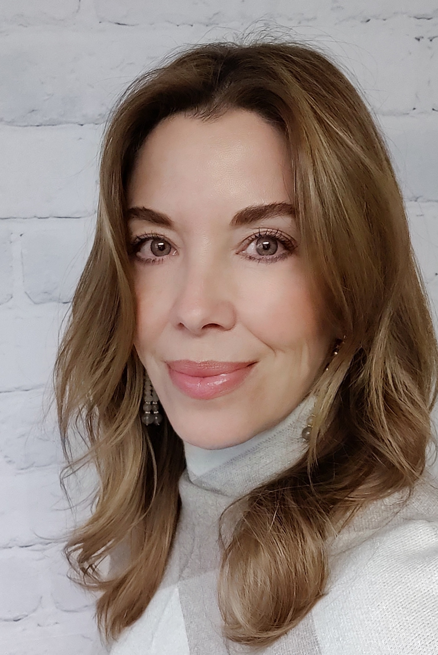 Tina with the long brown hair and earrings smiling in front of a white brick wall