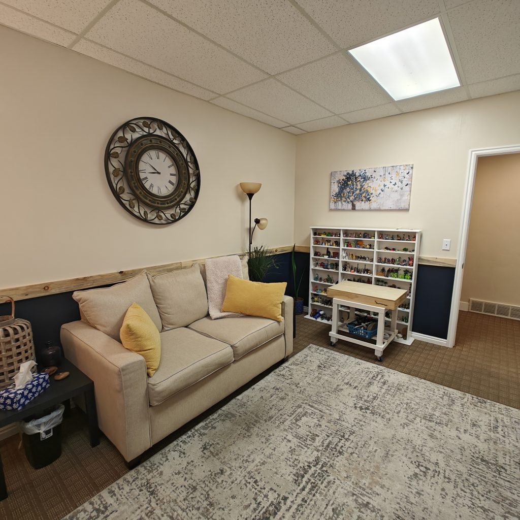 Cozy living room with beige sofa, yellow pillows, wall clock, and toy shelves in a well-lit space