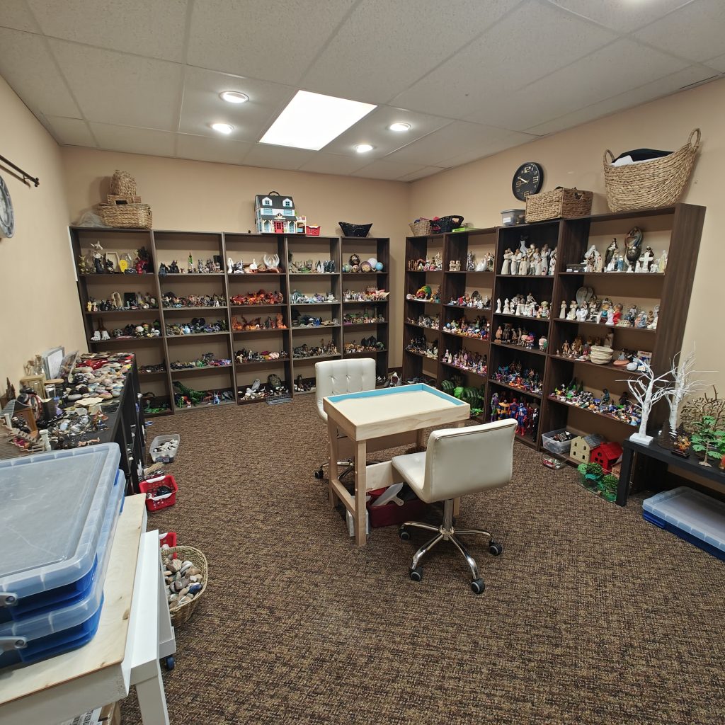 Room filled with shelves of figurines and toys, featuring a central table and chairs for creative activities